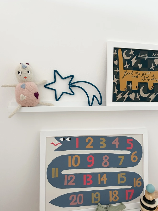 A shooting star shaped wall sign made from wire and knitted cotton yarn on a photo shelf in a kids room with other children's rooms decorations and wall prints. 