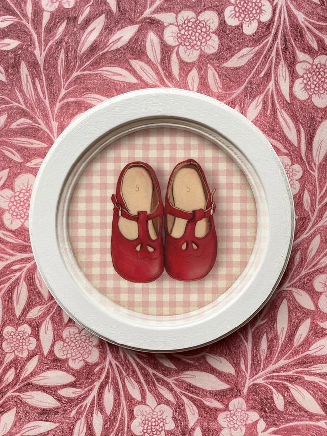 a photograph ofa framed illustration of a pair of red baby shoes in a beautiful ivory round wooden frame laid on illustrated wrapping paper