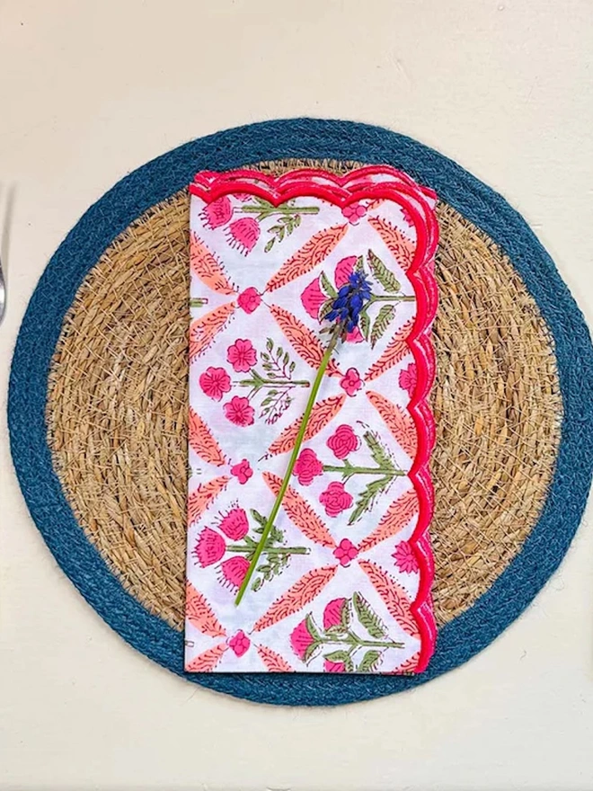 Close up of a round woven seagrass Placemats with teal rings, shown on a light surface with a flowery napkin placed on top. 