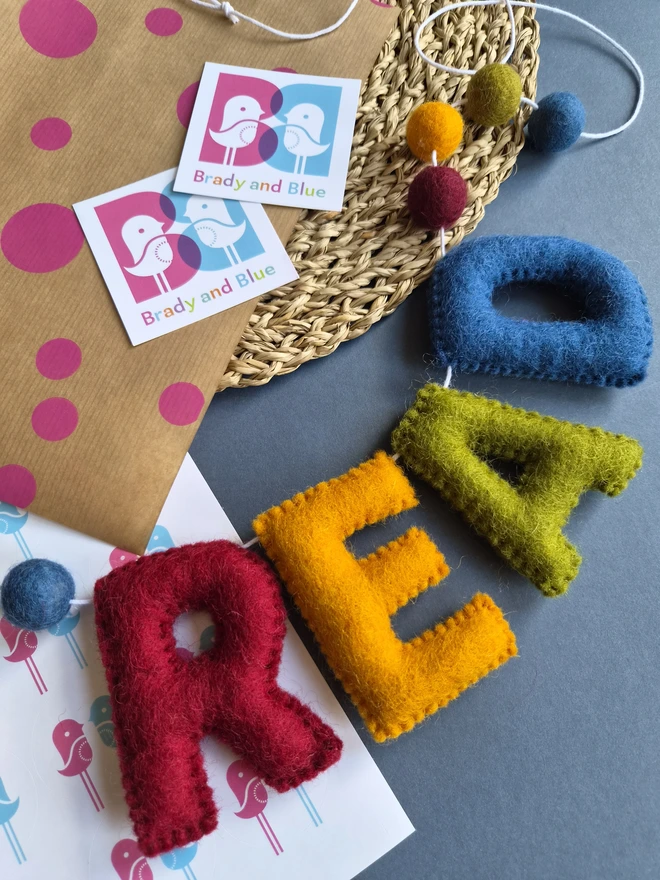 A garland in rich vintage rainbow colours laid on a table with some packaging. The garland consists of the letters READ and some felt balls.