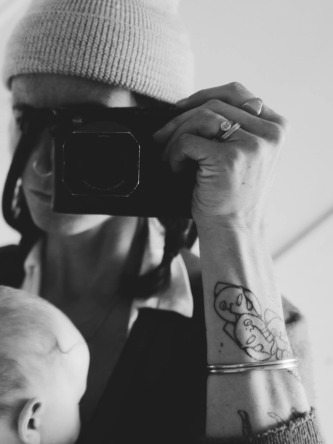 Black and white Image of mother taking her own photo in the mirror, with her baby on her chest, and holding her camera in her hand. She is wearing 2 bangles, and 3 rings, one ring has a small moon face 