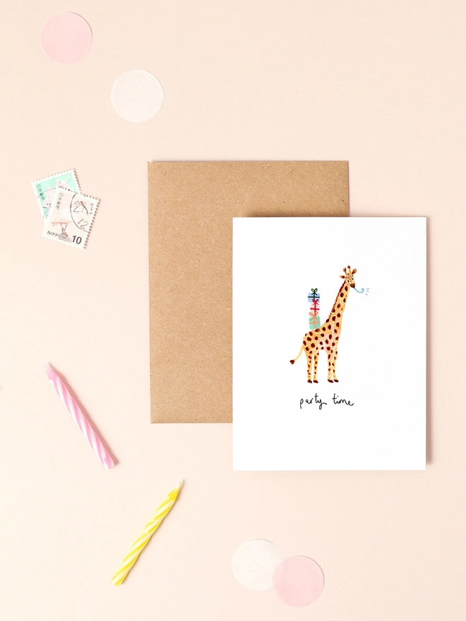 An image of a mini, rectangular white greetings card with an illustration of a giraffe with a stack of three colourful presents perched on it's back. A hand written 'party time' message is seen under the central illustration in black ink. The mini greetings card is placed on top of a rectangular brown kraft envelope.