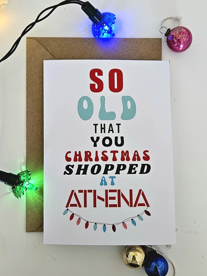 A photograph of the front of a humorous A6 greeting card illustrated using brightly coloured text and the retro shop logo. The text says so old  that you Christmas shopped at Athena with string lights