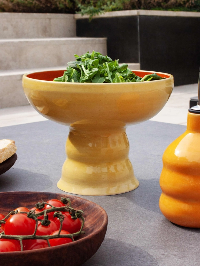 a colourful pedestal bowl filled with salad. the bowl has a wide shallow bowl and a chubby elevated foot. The bowl is butter yellow on the outside and tangerine orange on the inside. The bowl is sitting on a patio table surrounded by a tablecloth and matching tumblers