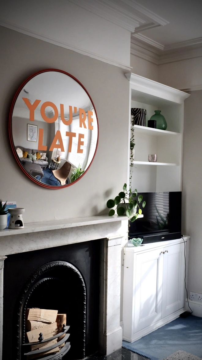 Round convex mirror with 'You're Late' handpainted in orange in a red round frame, above a cream fireplace surrounded by house plants