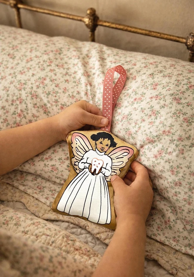 A child's hand gently holding a black afro carribean tooth fairy angel decoration on a child's bed. the tooth angel is holding a small ceramic tooth button, she is made from fabric and has a ribbon for hanging