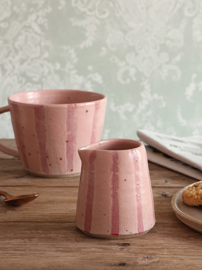 Handmade Ceramic Stoneware Small Jug in Pink Stripe sat on a wooden surface. A mug in the same style and a newspaper sit behind a mug, while a plate of cookies can be seen to the right. 
