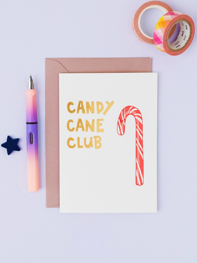 White Christmas greeting card featuring a hand-drawn red and white striped candy cane with gold foil Candy Cane Club lettering, displayed with a kraft envelope.