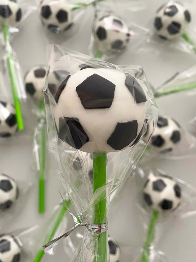 Football Cake Pops (Pack Of 6, 12, 18, 24), football cake pop placed into a cellophane bag in front of a background of more cake pops. This is against a white surface. 