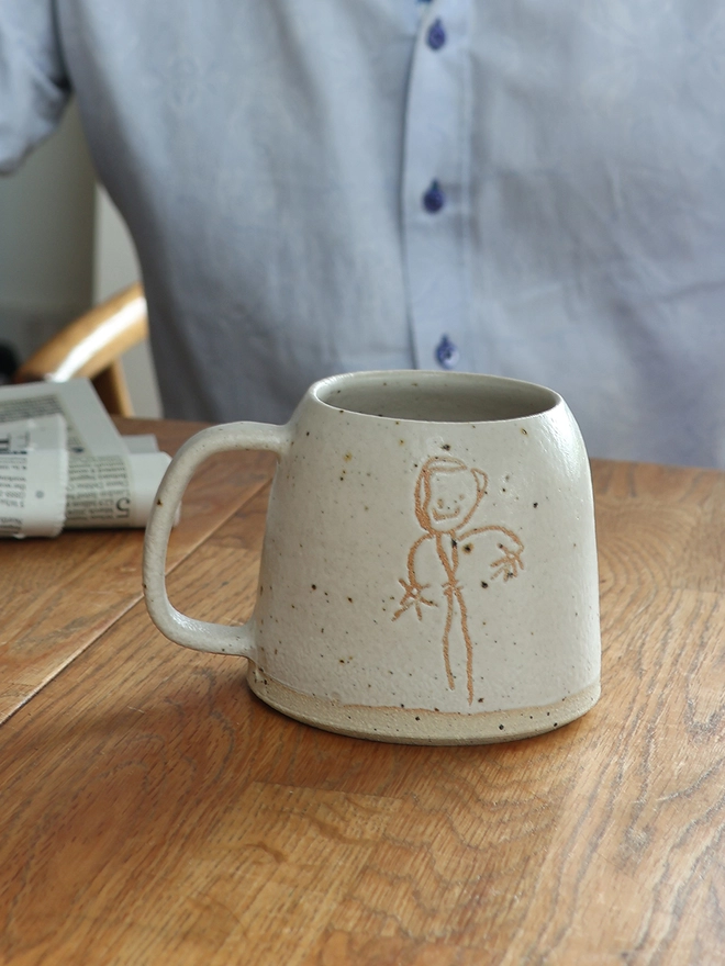 Childrens Drawing Handmade Ceramic Mug on a wooden table with a man in the background. The drawing features a stick man figure. 