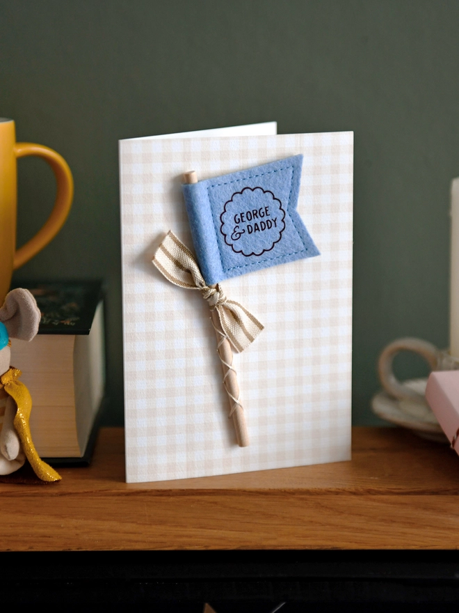 Personalised Father’s Day card with soft blue felt flag reading “George & Daddy”, handmade keepsake card with ribbon and wooden stick detail. The card stands on a wooden mantle beside a book, mug, candle, and wrapped gift.