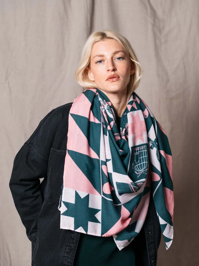 A model wears a large light weight forest green and calamine scarf displaying a range of motifs representing iconic women 