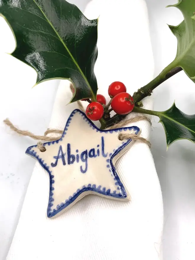 Personalised Ceramic Christmas Star Name Place Markers, a colourful ceramic start sitting on a white surface attached to a holly bush. 