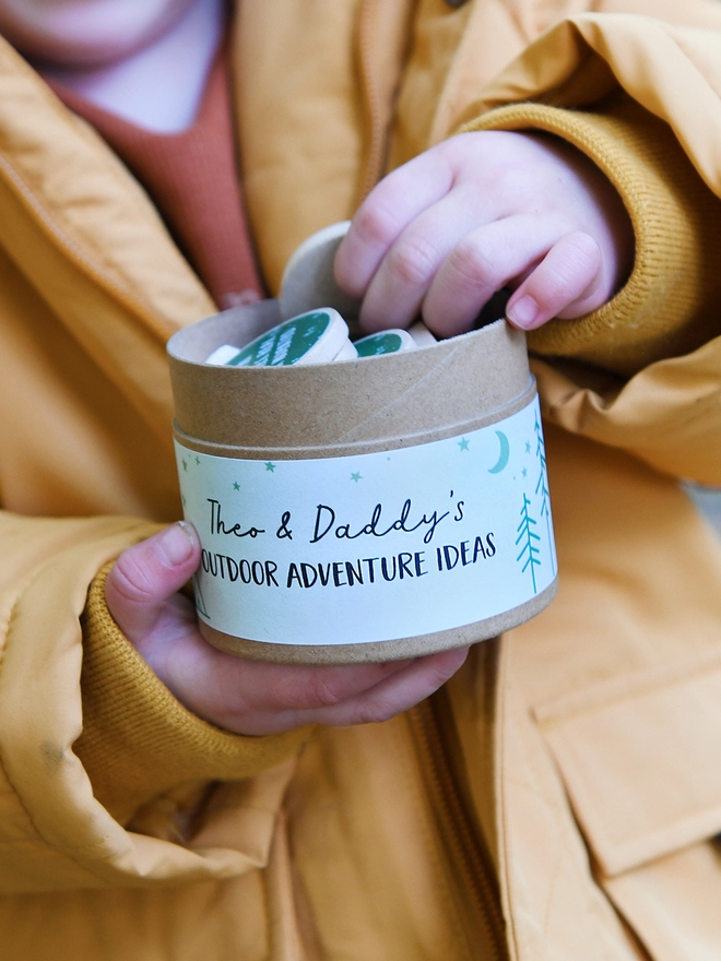 A young child wearing a mustard yellow coat holds a cardboard jar with several wooden tokens poking out of the top. A label on the outside reads Outdoor Adventure Ideas.