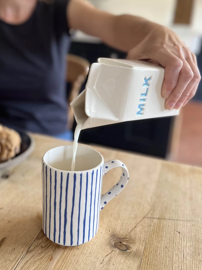 A girl is pouring milk into a mug using handmade ceramic jug, that emulates a paper carton.