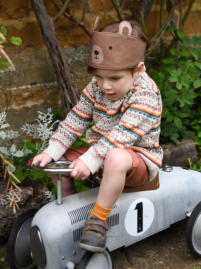 A young child wearing a knitted jumper and a bear dress up fabric crown sits on a ride on vintage car in the garden.