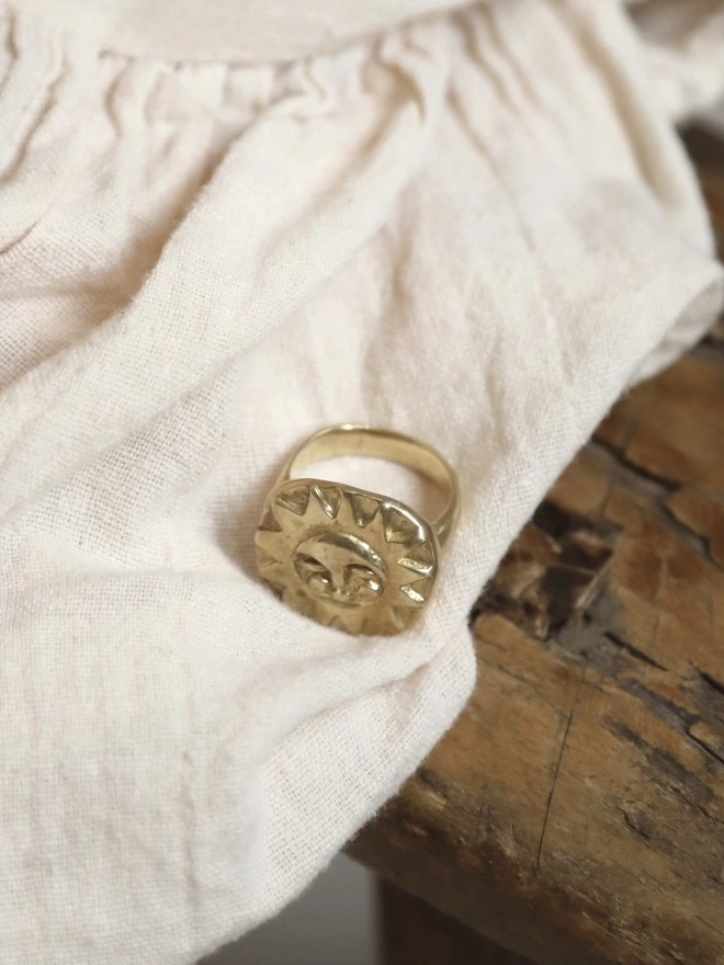 An image of a hand carved gold tone brass sun face ring on some cream linen type fabric and a piece of rustic wood 