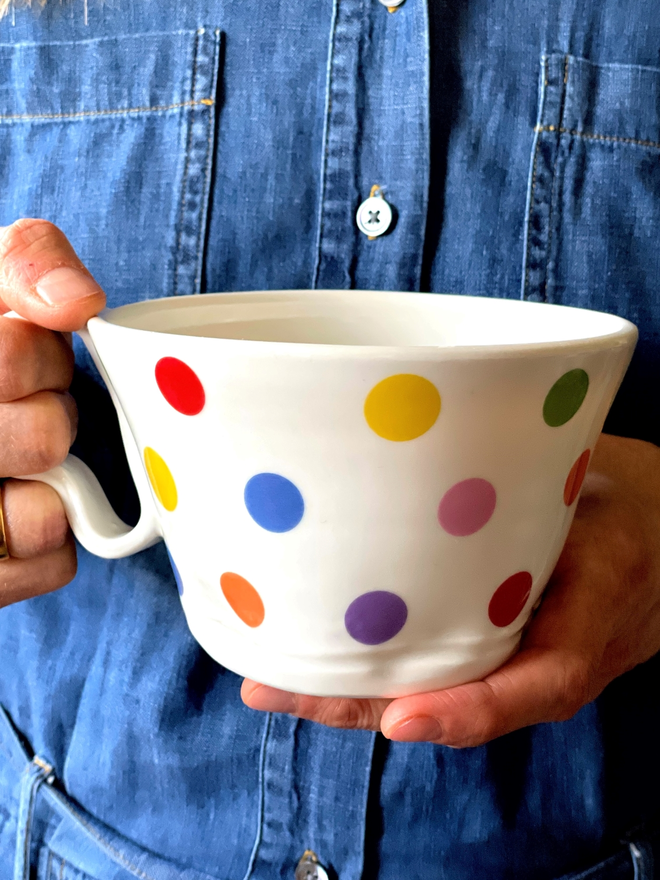 person holding a large porcelain cup with yellow orange blue green pink purple and red polkadots on a denim blue background