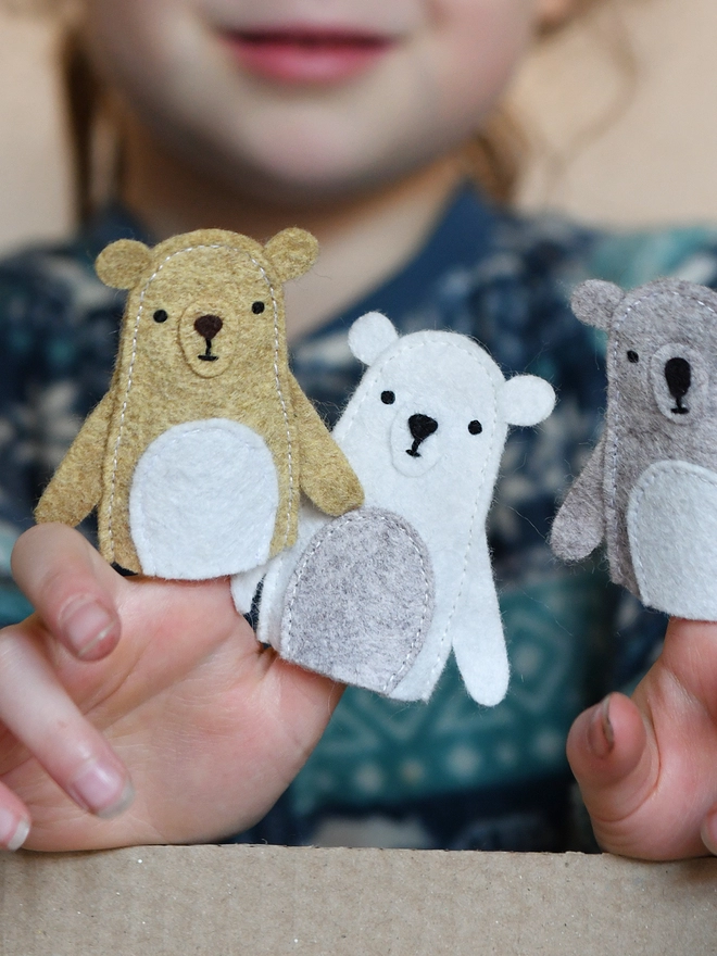 A young child wearing a blue jumper holds three handmade felt bear finger puppets on their fingers.