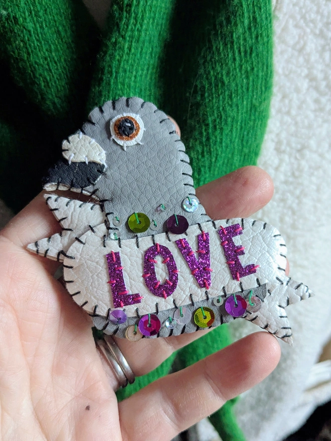Hand-stitched white and grey faux leather pigeon brooch with purple glitter ‘LOVE’ lettering and colourful sequin details, pinned to a bright green scarf