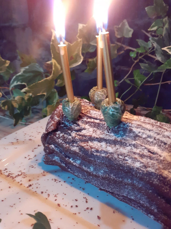 Three handmade ceramic Brussels sprout candle holders with lit gold candles decorating a chocolate Christmas log cake, surrounded by festive greenery.