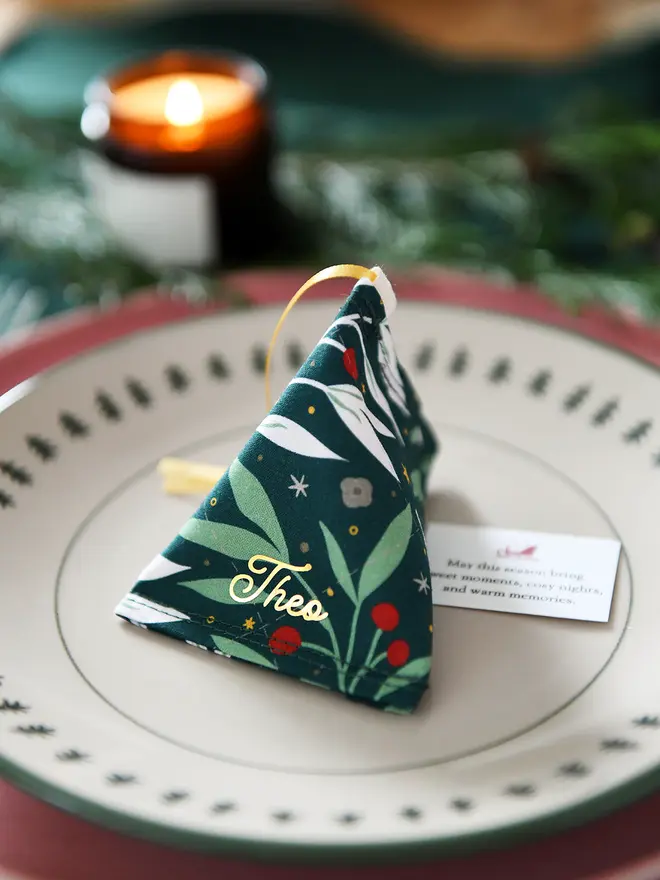 A green festive foliage fabric fortune cookie with gold personalised name “Theo” sits on a Christmas dinner plate, ready as a handmade festive place setting with a hidden Christmas wish card beneath.