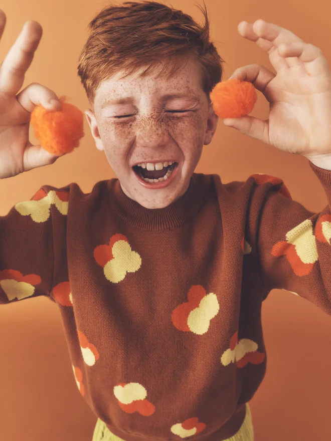 Close crop of a child laughing while holding an orange pom-pom in each hand, wearing a Studio Koter chestnut jacquard jumper.
