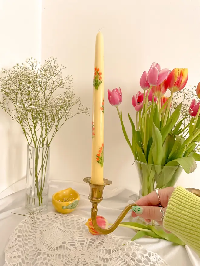 Mon Cherry & Tulip Fever Candle, a woman placing the candle on a table set accompanied by flowers in vases. It's floral design wraps around the stalk. 