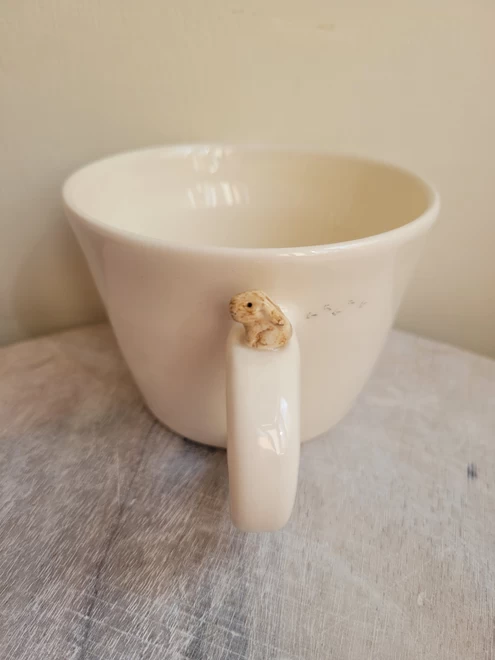 White ceramic mug with a tiny beige model bunny on the handle and small foot prints on the cup it is on a pale wooden board