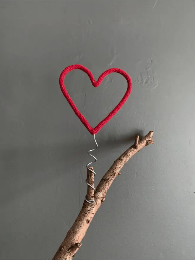 A red string wrapped wire heart shaped Christmas tree topper on a branch against a grey background