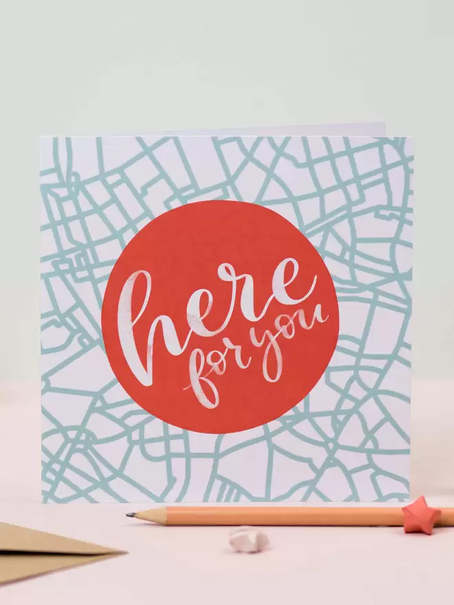 A square card features a red circle that says 'here for you' in white lettering. The background of the card is an abstract map design in duck egg blue.