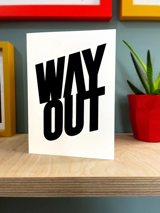 WAY OUT screenprinted in black, with an arrow pointing the way, embedding in the negative space of the lettering. Sat on a plywood shelf, duck egg blue background, with glimpses of picture frames around a bit of a plant showing.