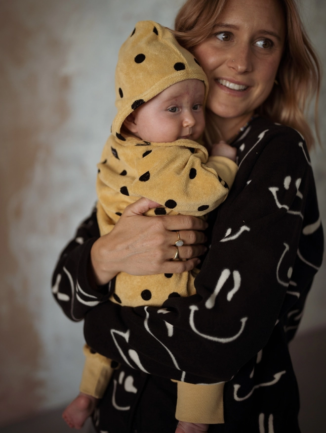Baby wearing the Camel Terry Towel Spot Baby Sleepsuit held by a woman.