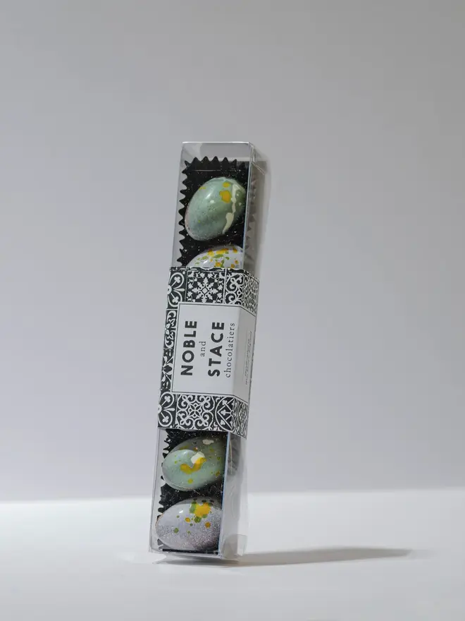 Clear rectangular box filled with assorted solid milk and dark chocolate mini eggs in pastel colours with speckled paint-style decoration, displayed on a white background.