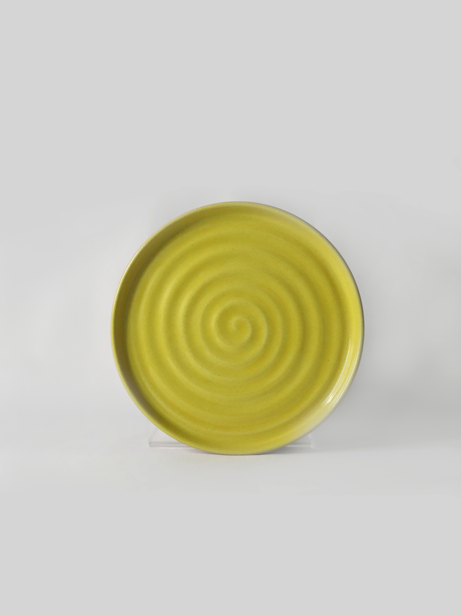 A handmade ceramic cake plate in sun yellow glaze, photographed from the front against a plain light grey background, showing a natural concentric swirl texture and a shallow raised rim