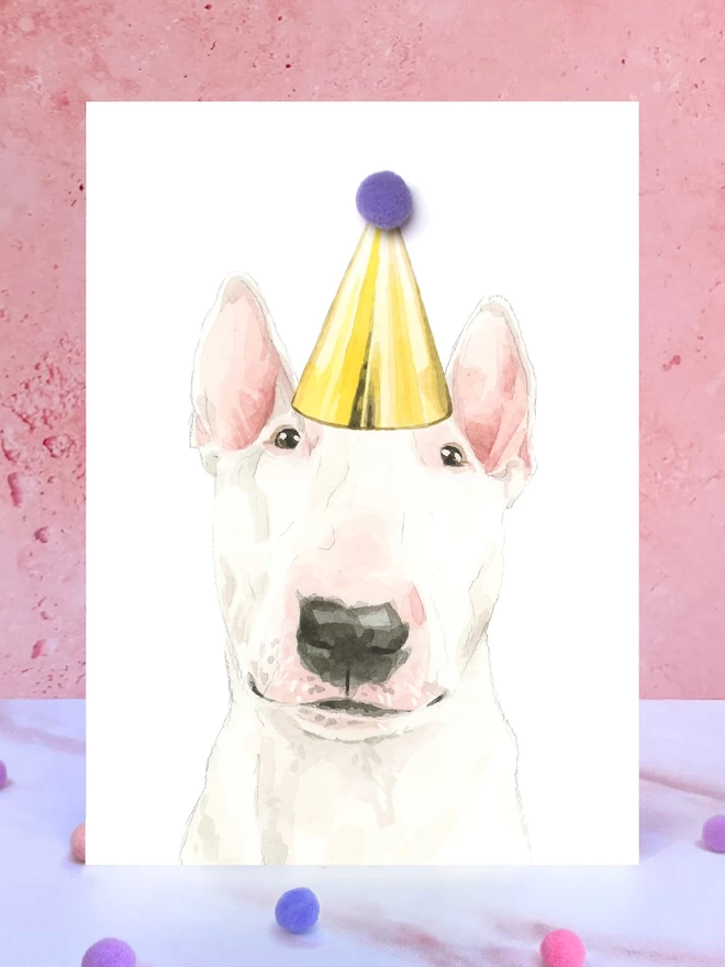 A greeting card featuring a hand painted design of a english bull terrier, stood upright on a marble surface surrounded by pompoms. 