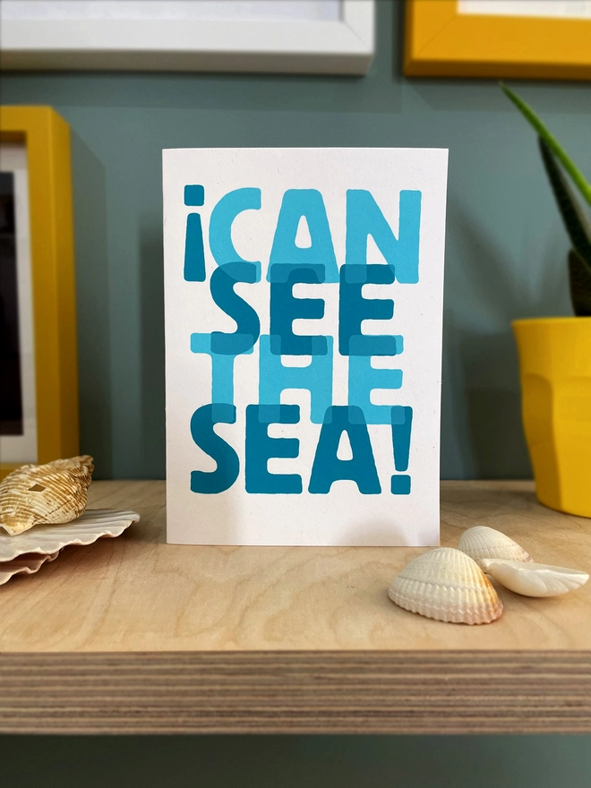 I can see the sea, screenprinted words overlapped in two turquoise blues, on a white card. It is sat on a plywood shelf, with shells, edges of picture frames and a plant half in shot.