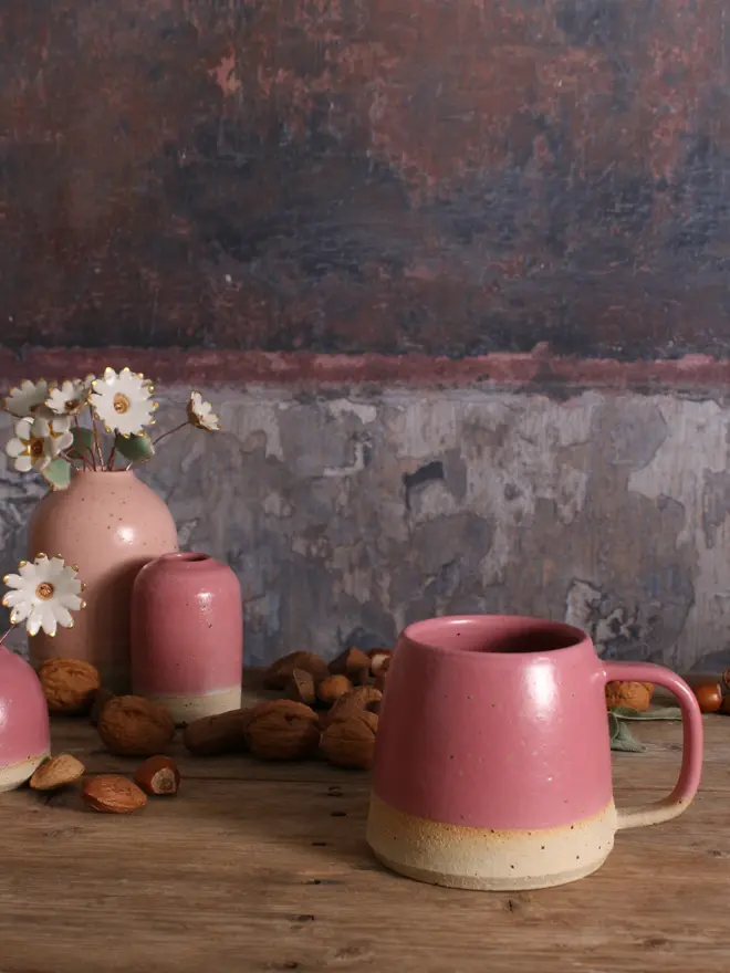 Ceramic Fuchsia mug sat on a wooden surface. The background shows a pale pink ceramic vase holding a ceramic flower bouquet alongside a small ceramic fuchsia vase.