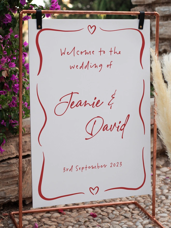 a white board hanging from some black leather straps from a copper pipe frame, on a cobbled path in front of a wall with some purple flowers and green bushes. the sign is in red writing and says welcome to the wedding of Jeanie and David on the 3rd September 2023