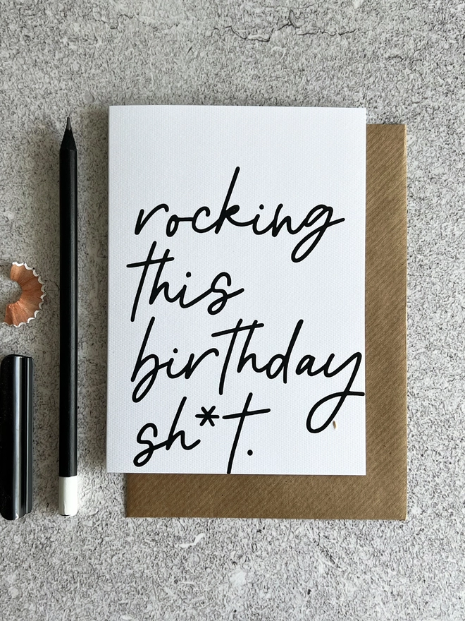 White card that reads "Rocking this birthday sh*t" in cursive, laid on a brown envelope next to a black pencil.