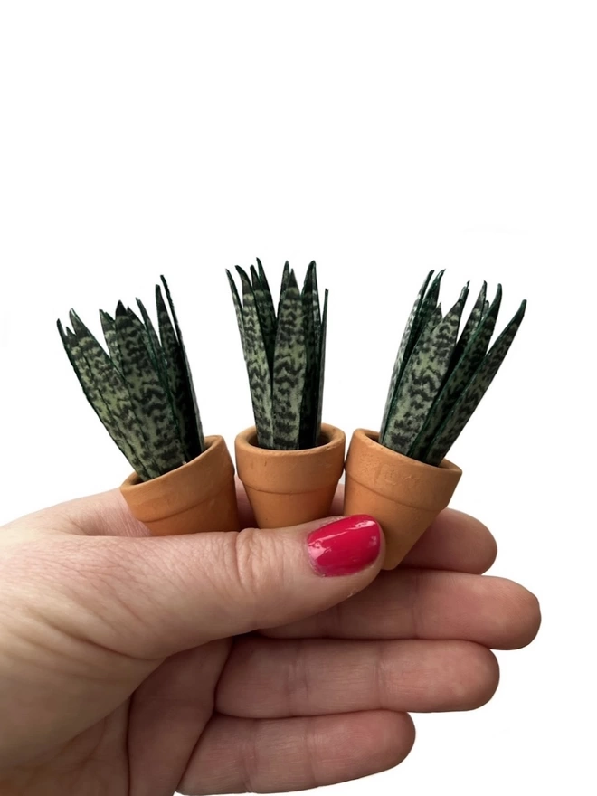 3 miniature replica Sanseveria Snake Plant paper plant ornaments in terracotta pots being held in one hand against a white background