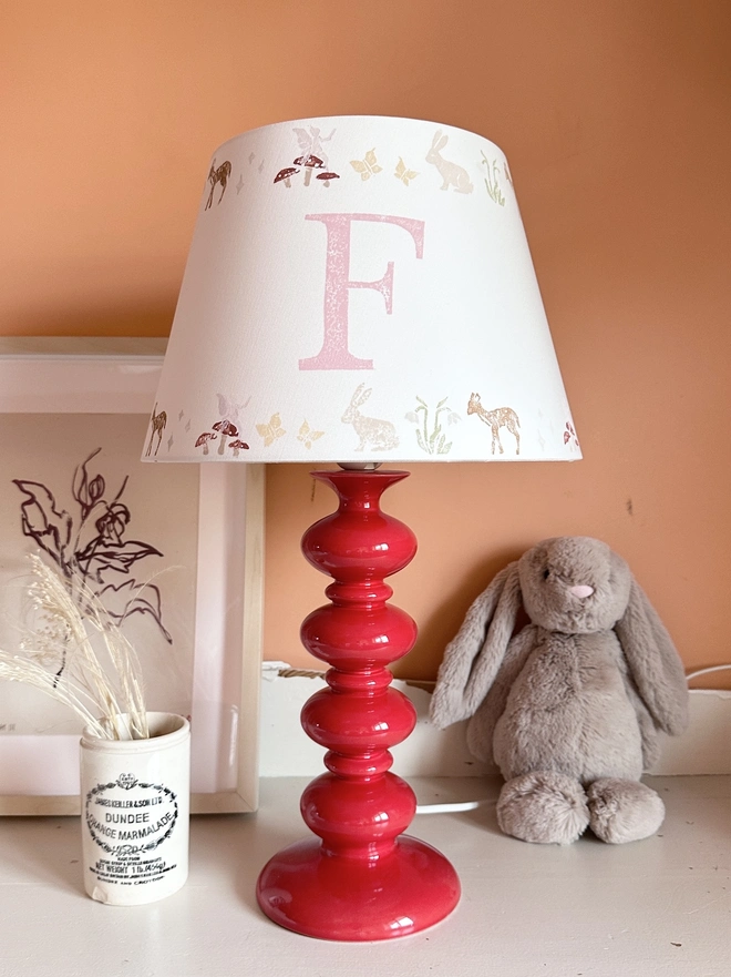 Personalised kids lampshade with woodland animals design and pink initial and date, styled on a red lamp base in a girl’s bedroom. A pretty empire lampshade ideal for kids bedroom décor and new parent gifts.