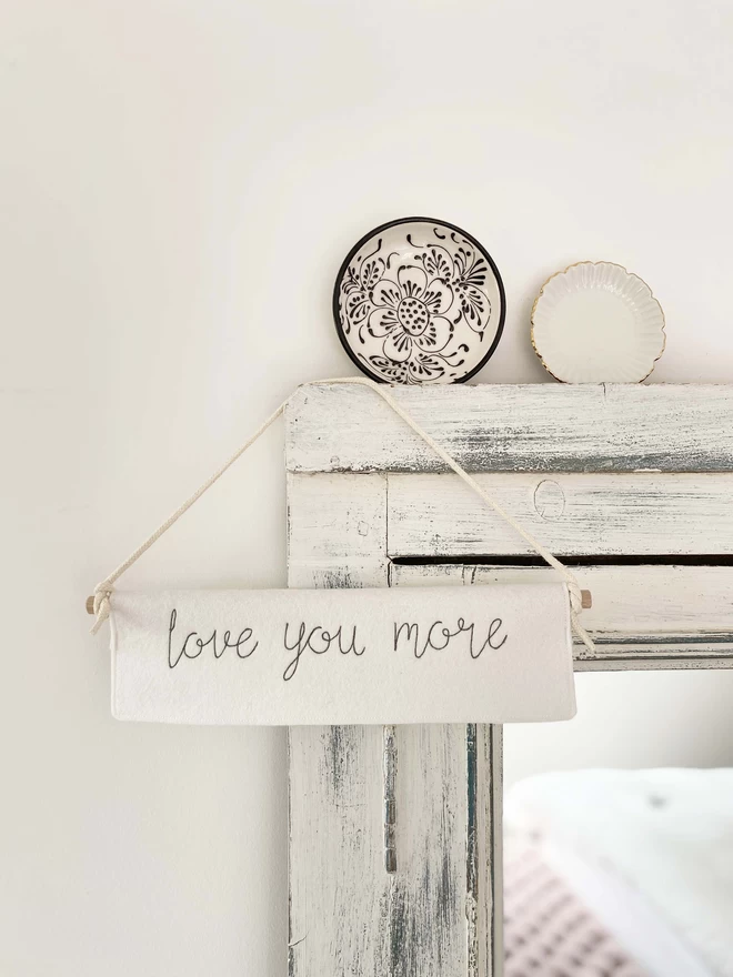 Love You More Felt Embroidered Banner hanging on mirror with plates
