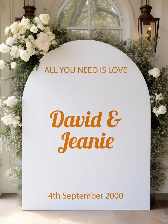 An board in the shape of an arch, white background with the words all you need is love, David and Jeanie and the date of the wedding in orange lettering, it is leaning against a window with white flowers, foliage and lanterns surronding it 