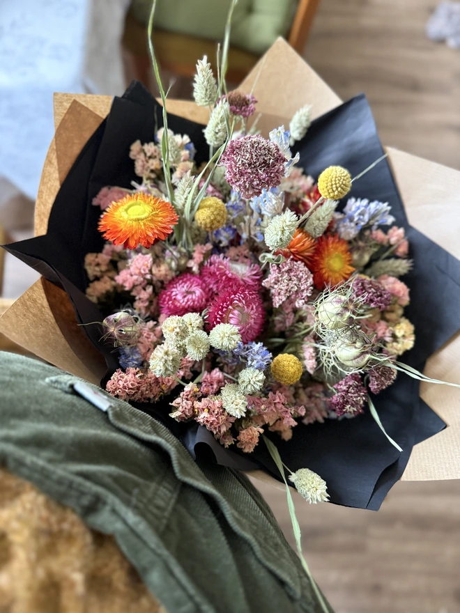 a dried flower bouquet in bright colours wrapped in black tissue and kraft paper held by a person wearing a mustard coloured jumper and green dungarees