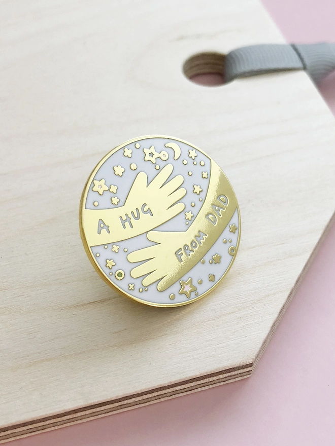 A white and gold pin badge with a hugging arms design and the words “A hug from Dad” is placed on a wooden tag.