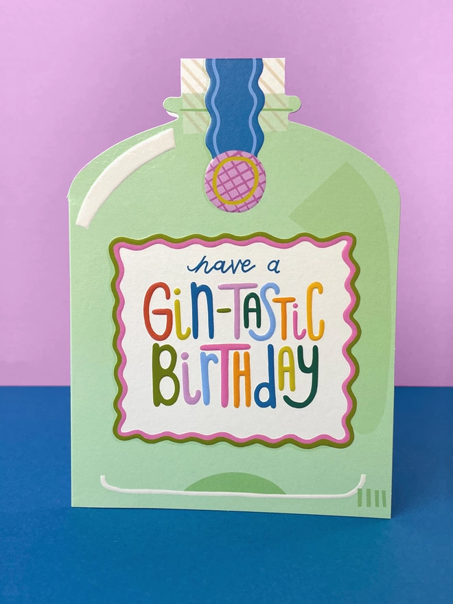 A vibrant die cut birthday card in the shape of a mint gin bottle with a rainbow coloured ‘Have a gin-tastic birthday’ message