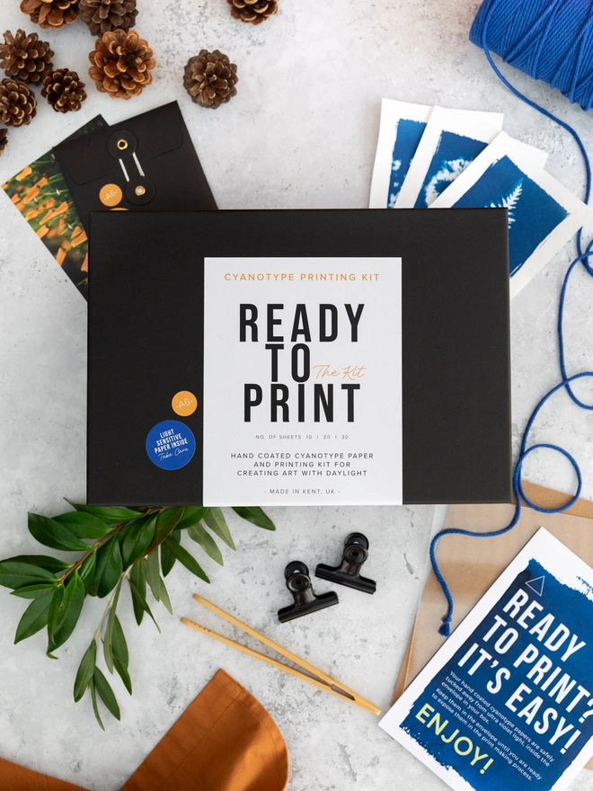 "ready to print" black cyanotype printing kit box displayed with contents and finished cyanotype prints.