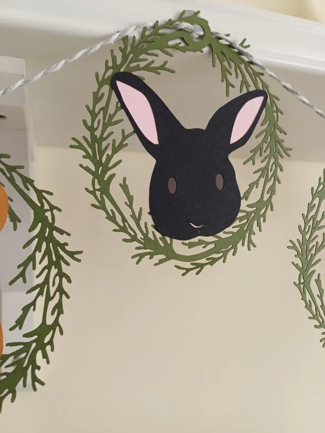 Closeup of bunny decoration. Bunny has black face, dark brown eyes and pink ears. Surrounded by lasercut olive green foliage oval frame