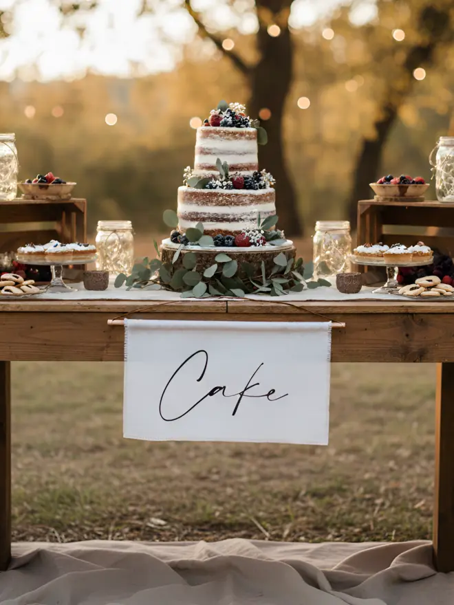 A linen cake banner hanging from a rustic wedding cake table at an outdoor ceremony